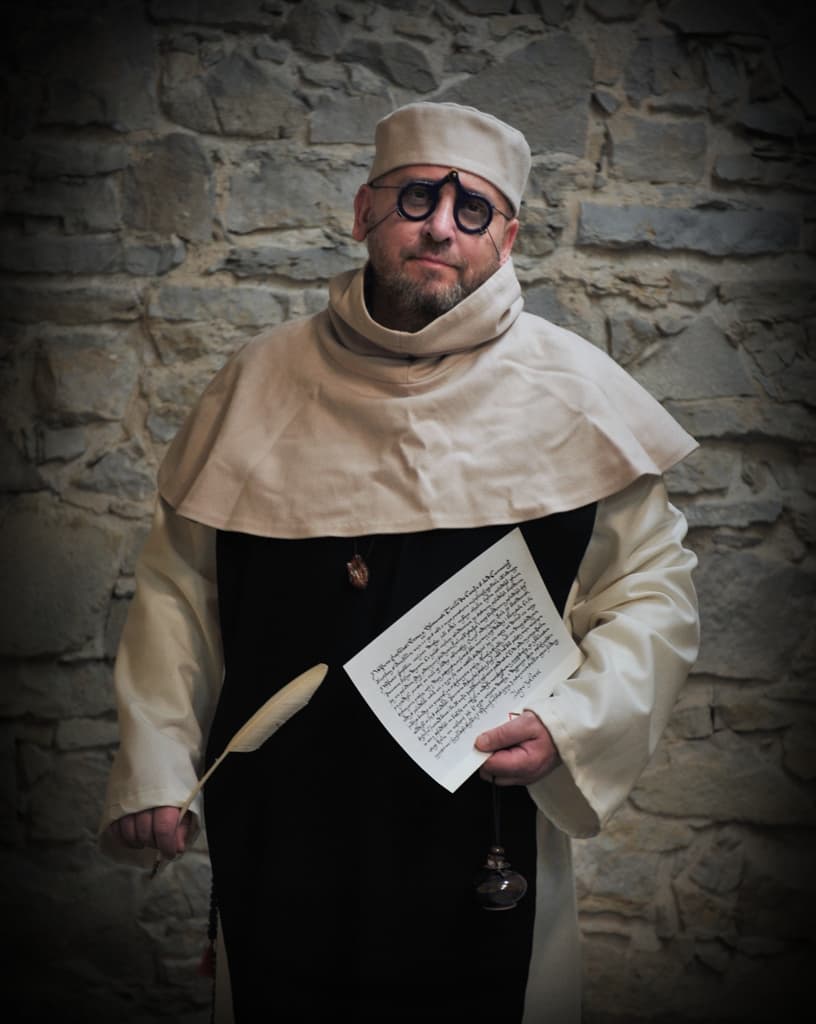 A man in a medieval-style costume with a cowl and cap, holding a quill and a handwritten letter, standing in front of a stone wall